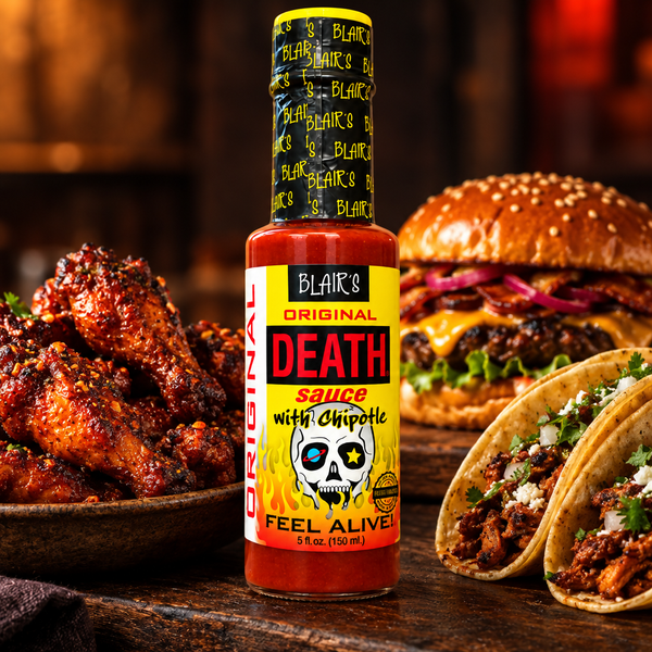 A 150ml bottle of Blair's Original Death Sauce with chipotle on a wooden tabletop surrounded by burgers, wings and tacos. Buy online at Deathsauce Australia.